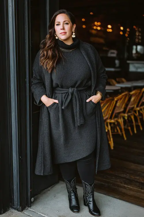 A plus-size woman wears a sweater midi dress layered over charcoal leggings with sleek black cowboy boots, finished with a belted wool coat and elegant statement earrings