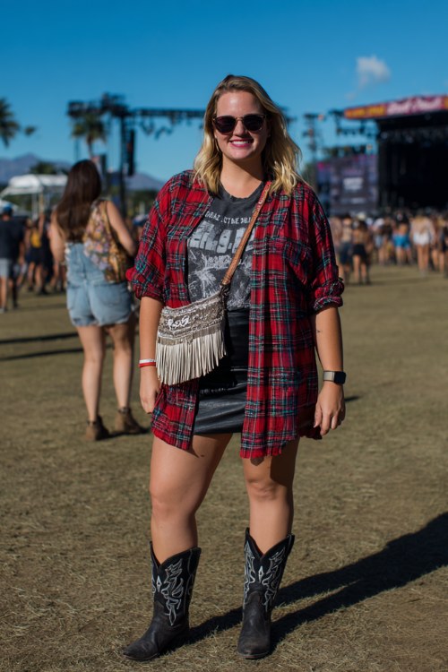 A plus-size woman wears black cowgirl boots with a faux-leather mini skirt, tucked-in bold concert tee and oversized plaid shirt worn open, finished with a fringe crossbody bag