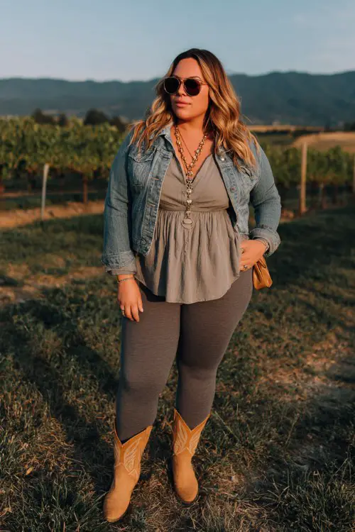A plus-size woman wears charcoal leggings with tan cowboy boots, a cropped denim jacket over a relaxed blouse, layered necklaces