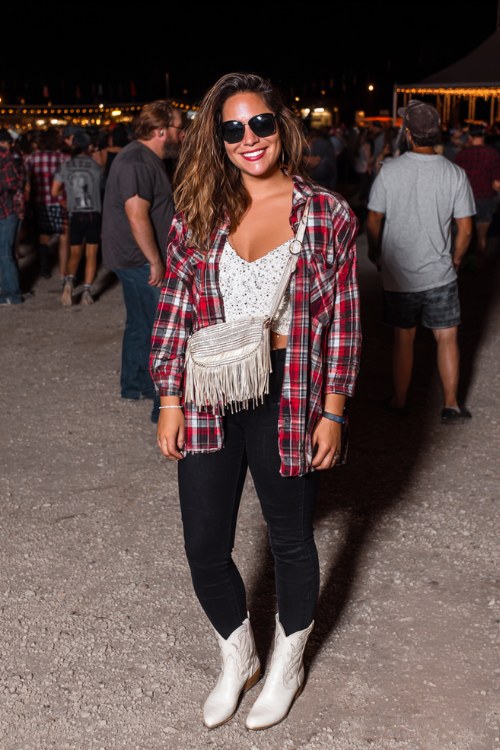 A plus-size woman wears white ankle cowboy boots with black skinny jeans, a sequin cami and oversized plaid shirt worn open, finished with a fringe crossbody bag and sunglasses