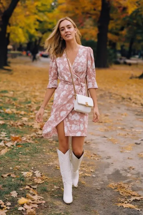 A woman over 30 wears a floral wrap dress in soft blush tones, paired with tall white cowboy boots and a crossbody bag
