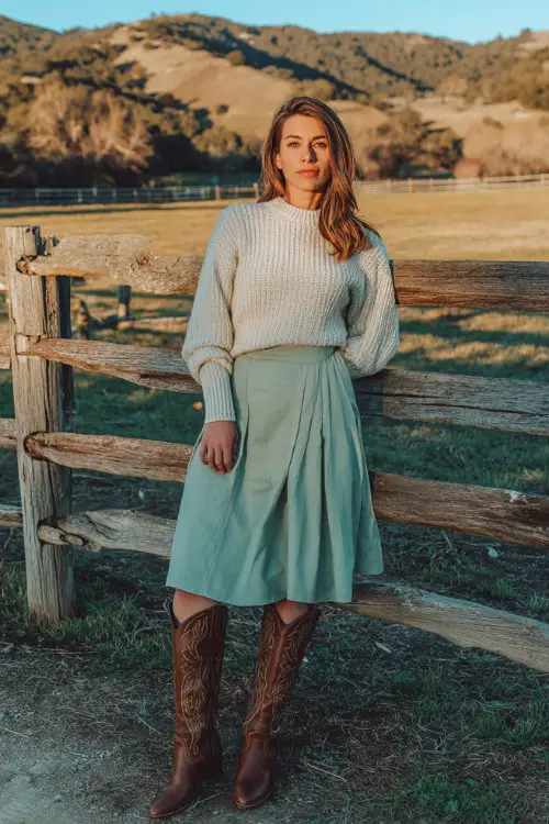 A woman over 30 wears a pastel green midi skirt with brown tall cowboy boots and a cream knit sweater