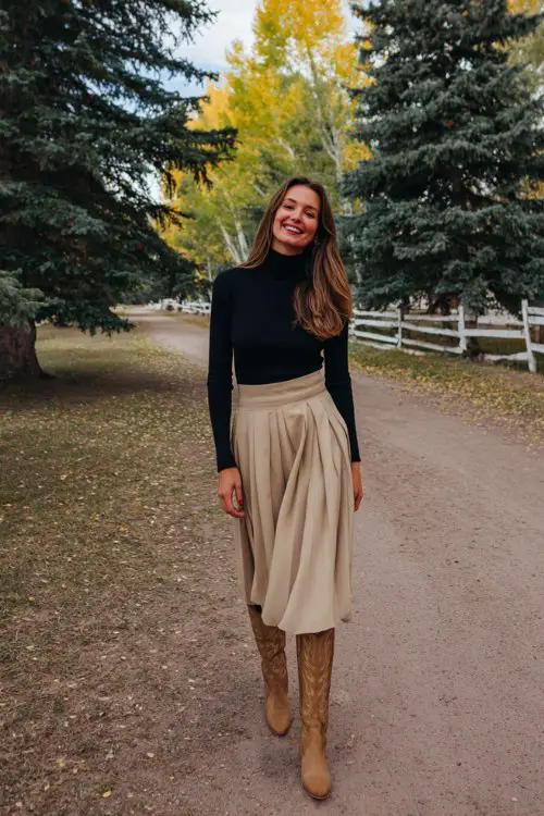 A woman over 30 wears a pleated beige midi skirt with tan tall cowboy boots and a tucked-in black turtleneck