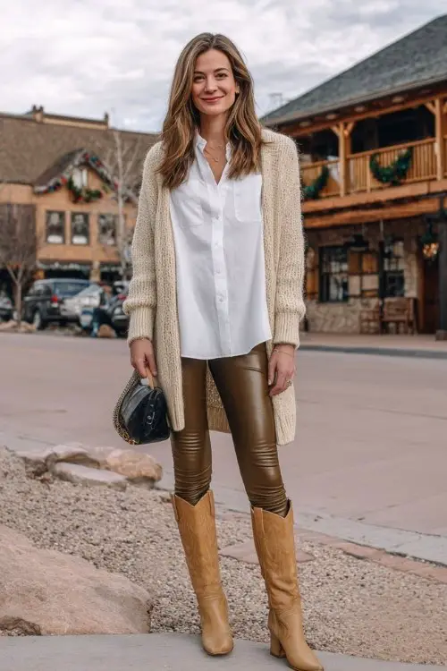 A woman over 30 wears faux-leather leggings with tan tall cowboy boots and a white button-down shirt layered under a cozy cardigan