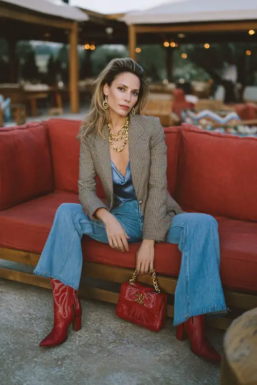 A woman over 30 wears red cowboy boots with flared blue jeans, a silky camisole and tailored suede blazer, accessorized with layered gold chains and a structured handbag