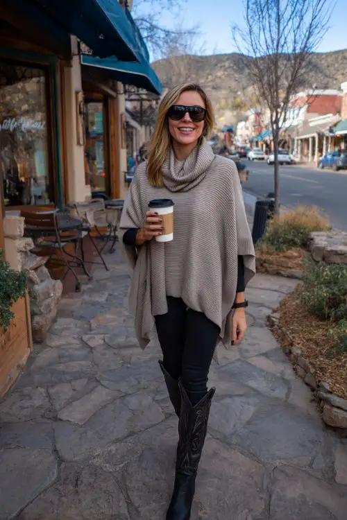 A woman over 40 wears black leggings with sleek tall cowboy boots and a soft cashmere poncho, holding a coffee cup while strolling through a cozy town street