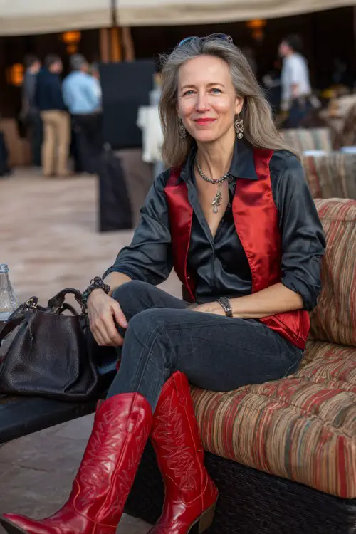A woman over 40 wears polished red cowboy boots with tailored dark jeans, a satin blouse and long statement vest, styled with elegant jewelry and a leather handbag