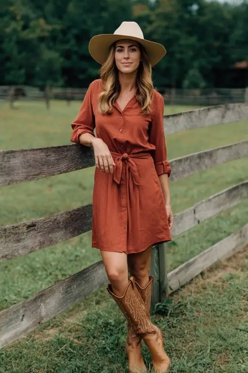A woman wears a rust-colored shirt dress with a tied waist, paired with tall tan cowboy boots and a wide-brim hat