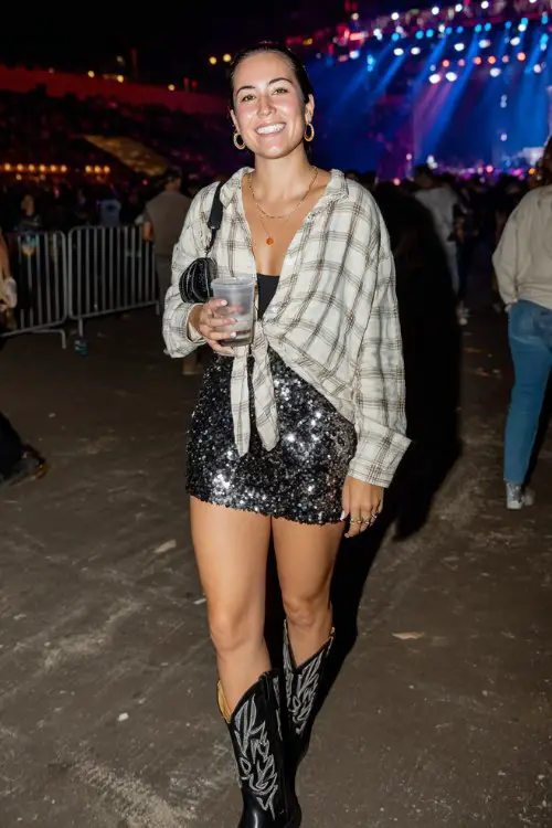 A woman wears black cowgirl boots with a sequin mini skirt and oversized flannel shirt tied at the waist, accessorized with hoop earrings and a sleek clutch