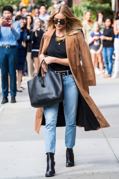 A woman wears black jeans with black ankle cowboy boots and a long camel coat