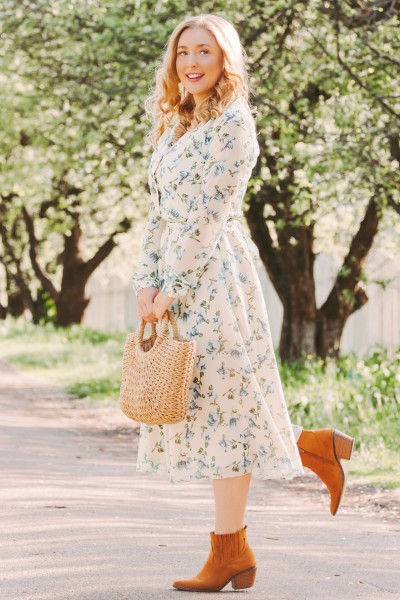 A woman wears floral dress with brown ankle cowboy boots and straw tote