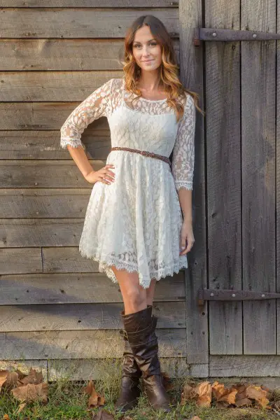 A woman wears lace dress and brown cowboy boots stand by the wooden house