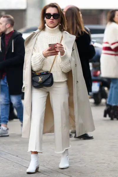 A woman wears white cowboy boots with a white jeans, sweater and long coat