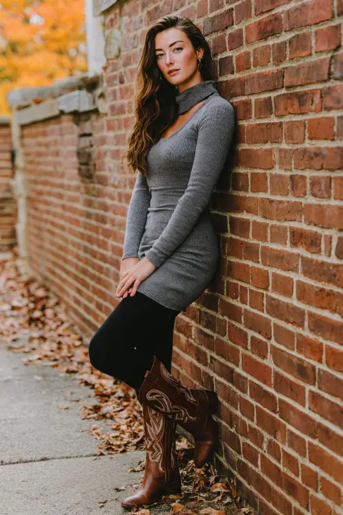 Brunette posing against brick wall with autumn leaves wearing black leggings with brown cowboy boots, fitted gray turtleneck sweater dress layered over leggings