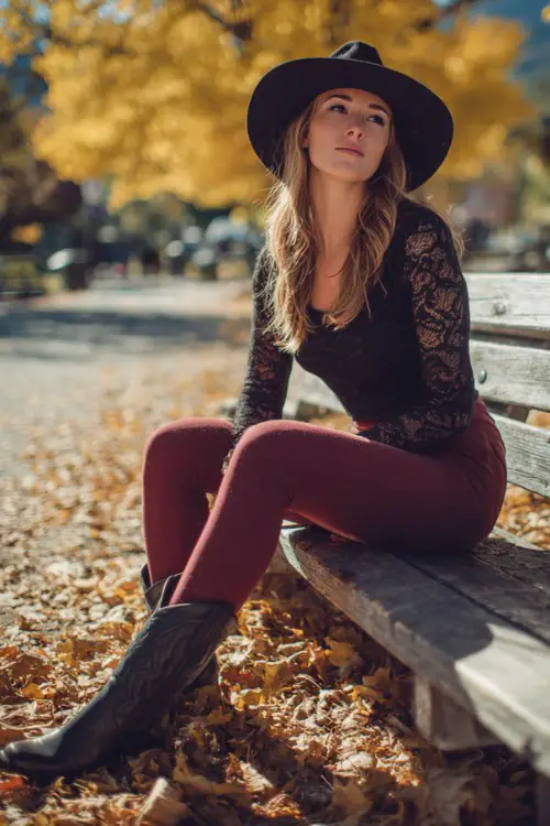 Woman sitting on park bench covered in fall leaves wearing burgundy leggings with black cowboy boots, fitted black lace top, wide-brim felt hat