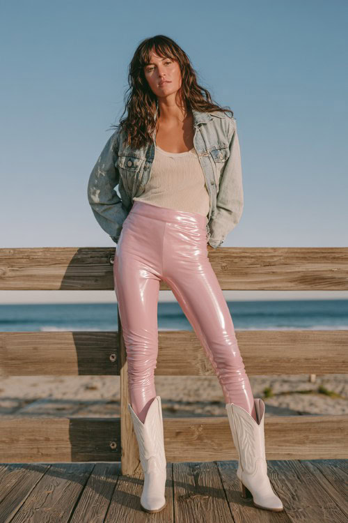 Woman standing on a wooden boardwalk wearing glossy blush pink leggings with white cowboy boots, light denim jacket over neutral top