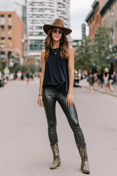 woman in a black sleeveless top with dark gray faux leather leggings and cowboy boots and western hat on the street