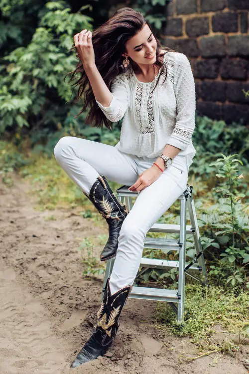 A woman wears black cowboy boots with white jeans and boho top