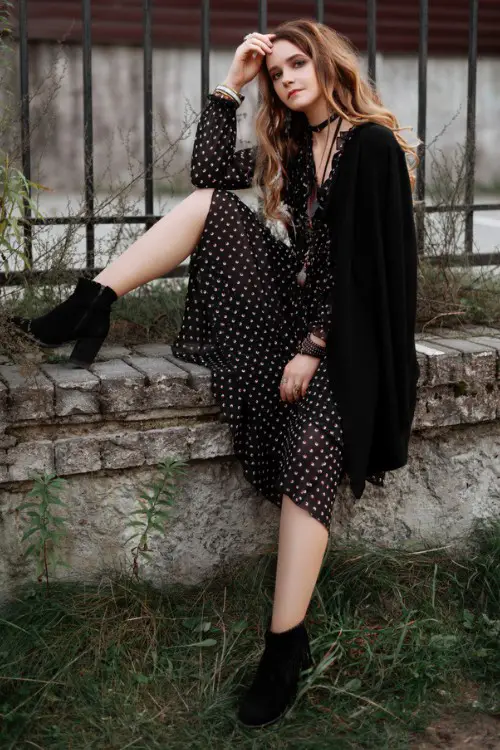 A woman wears black dotted dress, black cowboy boots and a black jacket