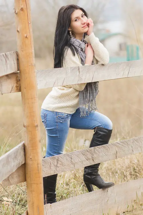 A woman wears cowboy boots with a sweater, a scarf and jeans