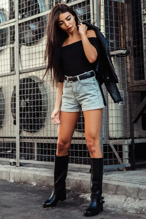 A woman wears cowboy boots with shorts, an off shoulder top and a leather coat