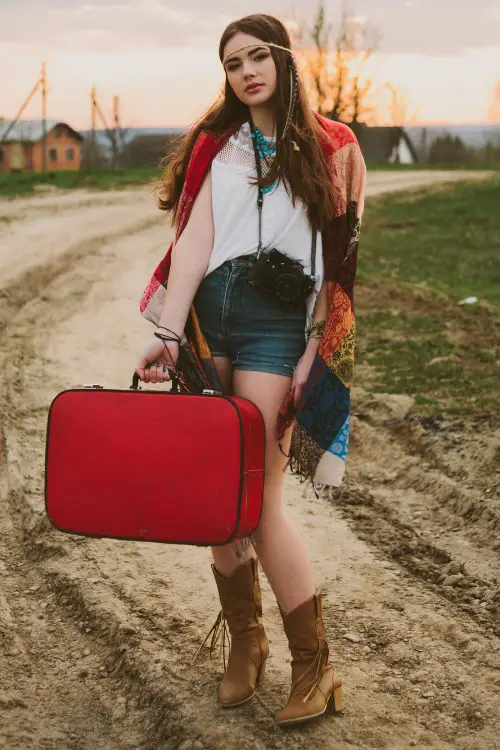 A woman wears cowboy boots with shorts and a boho shirt and a red bag