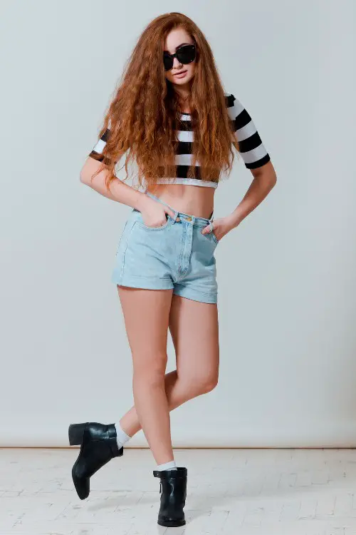 A woman wears cowboy boots with shorts and a striped shirt