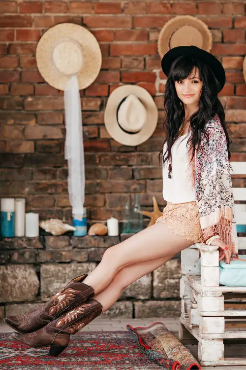 A woman wears lace short, white top, a coat and brown cowboy boots