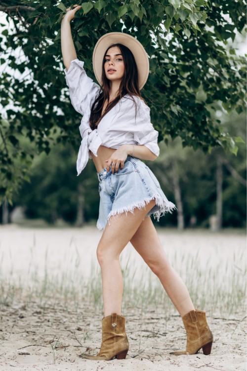 A woman wears ripped shorts with white top and brown cowboy boots