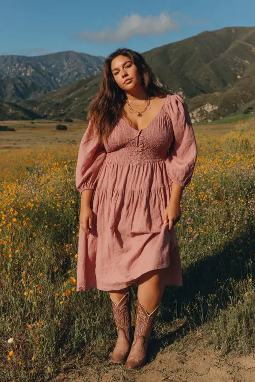 A curvy woman wears a smocked midi dress in dusty pink with puff sleeves, paired with embroidered cowboy boots and layered necklaces