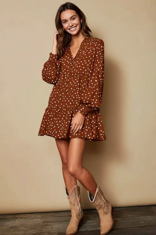 A woman wearing a loose brown polka dot mini dress with long sleeves and a flowy, tiered fit, styled with neutral-toned cowboy boots