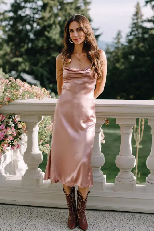 A woman wears a blush pink satin midi dress with a simple, elegant shape, styled with sleek cowboy boots and delicate accessories