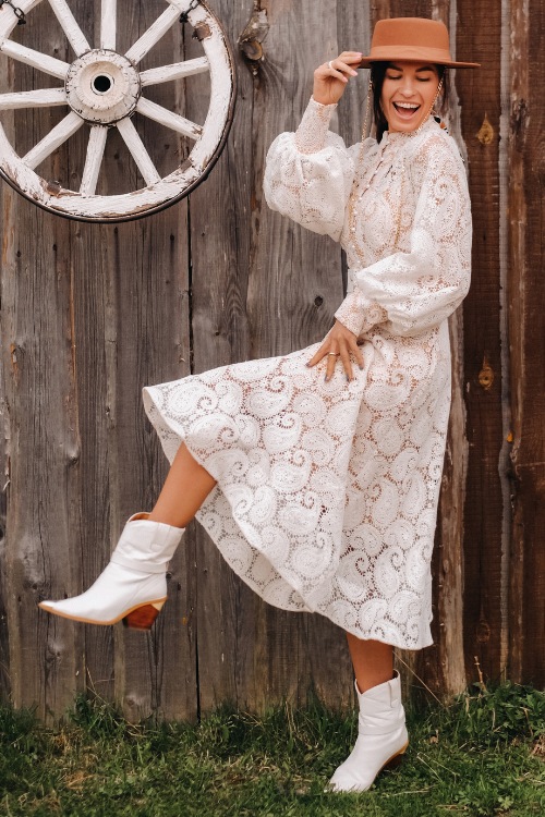 A woman wears a white dress with white cowboy boots and a brown hat