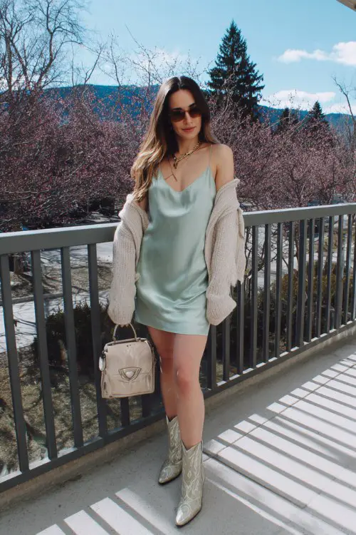 A woman wears a soft mint green satin slip dress layered with an oversized lightweight cardigan slipping off one shoulder