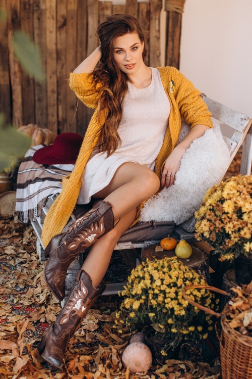 A woman wears brown cowboy boots with a white dress and a mustard coat