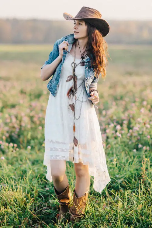 A woman wears cowboy boots with a long white dress and a denim coat