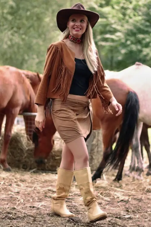 A woman wears cowboy boots with a short skirt, a black tank and fringe coat
