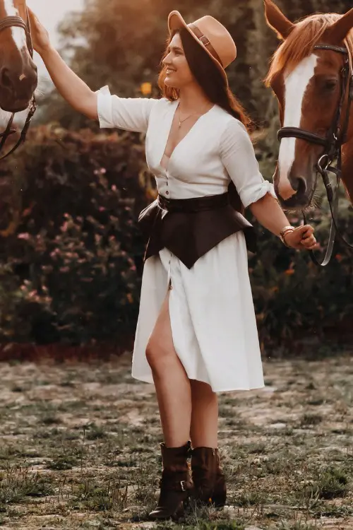 A woman wears cowboy boots with a white dress and a cowboy hat