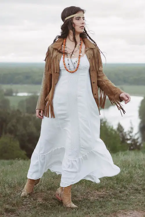A woman wears cowboy boots with a white dress and a fringe leather coat