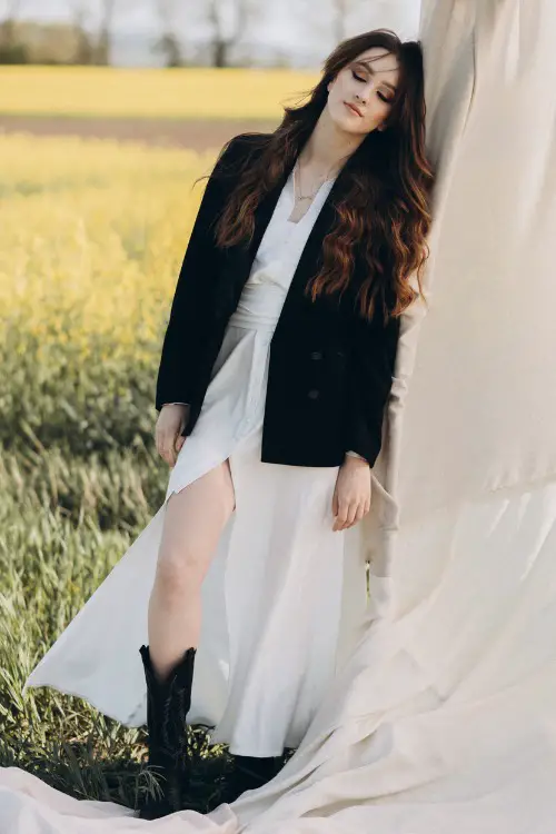 A woman wears a white dress, a blazer and black cowboy boots