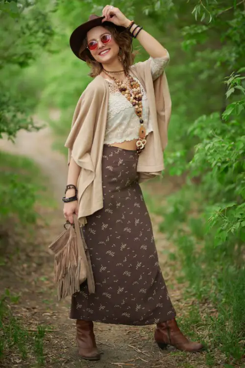 A woman wears long skirt, lace crop top, light coat and brown cowboy boots
