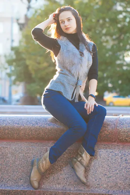 A woman wears long sleeves top, vest, jeans and a pair of cowboy boots