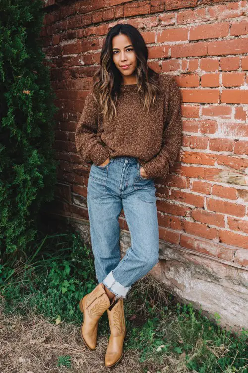 a woman wearing tan cowboy boots, patchwork blue jeans, and a cozy brown textured sweater