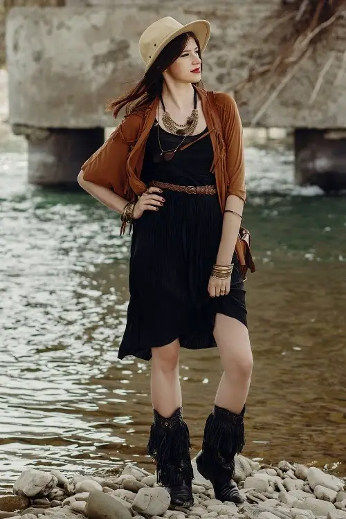 a woman wears black cowboy boots with black dress and suede fringe coat