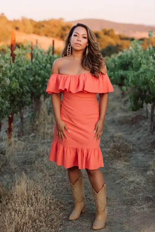 A curvy woman wears a bright coral off-shoulder midi dress with ruffled layers, styled with tan cowboy boots and gold hoop earrings