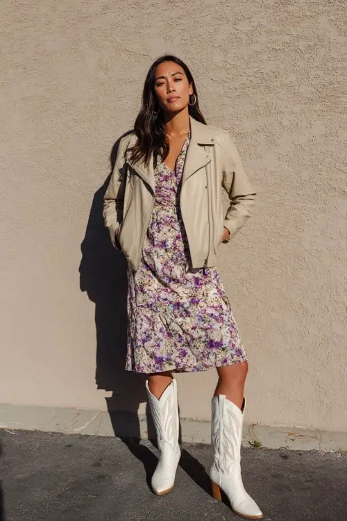 A woman in her 30s wears tall white cowboy boots with a floral midi dress and a beige leather jacket