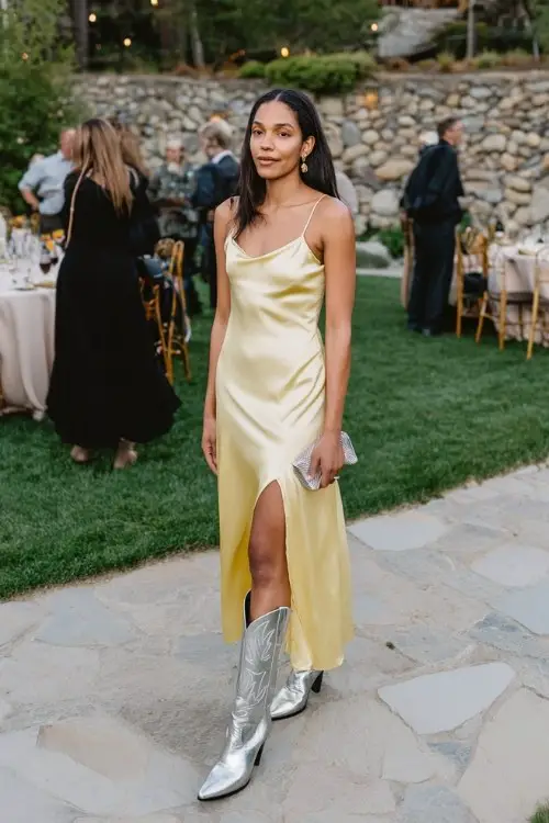 A woman over 30 wears a butter-yellow silk slip dress with a thigh slit, paired with metallic silver cowboy boots and a satin clutch
