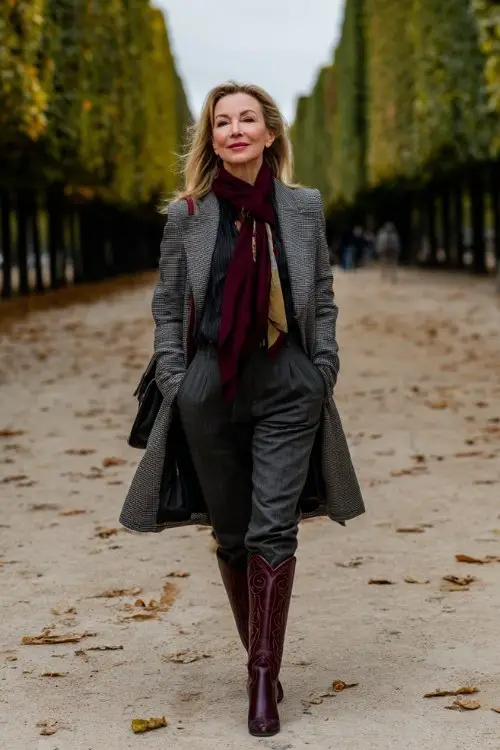 A woman over 50 wears tall burgundy cowboy boots, black tailored trousers, a gray wool coat, and a silk scarf