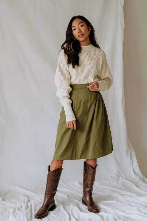 A woman wears tall brown cowboy boots, an olive green midi skirt, and a cream ribbed sweater