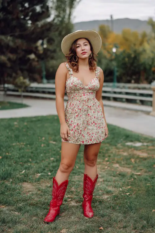 A curvy woman wears a floral mini dress styled with glossy red cowboy boots, hoop earrings and a straw hat